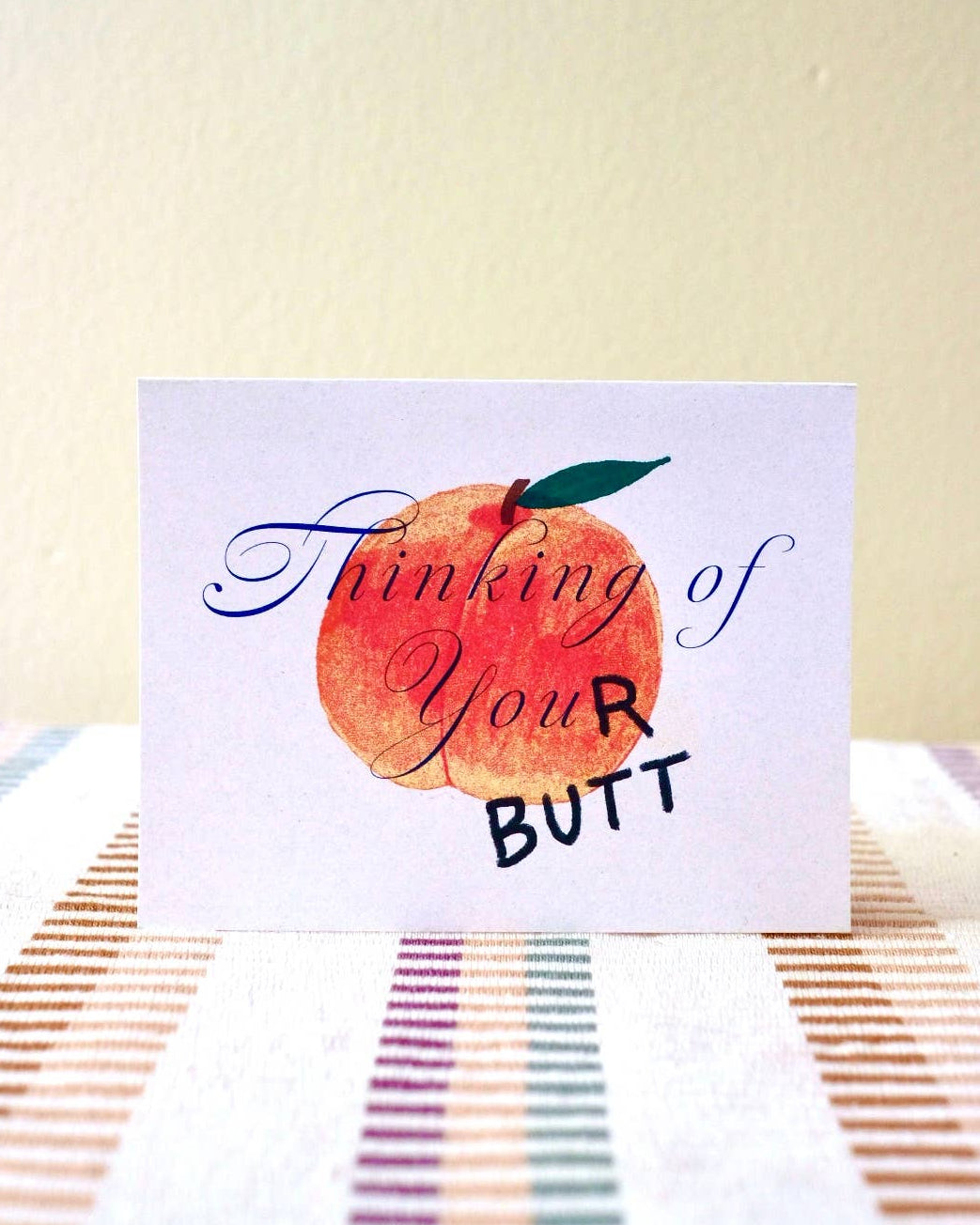 Card with a peach illustration and text that says "thinkig of your butt" on a striped surface